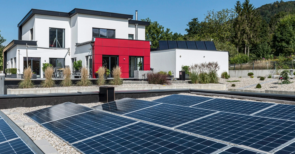 Moderne, zweistöckige weiße Haus mit rotem Fassadenteil auf einem Grundstück, umgeben von Bäumen. Auf dem Boden befinden sich Solarpaneele; im Garten sind Pflanzen und Gräser zu sehen.