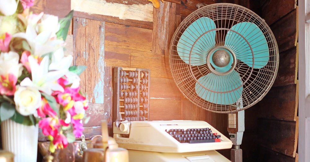 Ein großer Ventilator mit blauen Flügeln hängt an einer Holzwand. Daneben befindet sich eine Schreibmaschine und links ein Blumenstrauß in einer Vase.