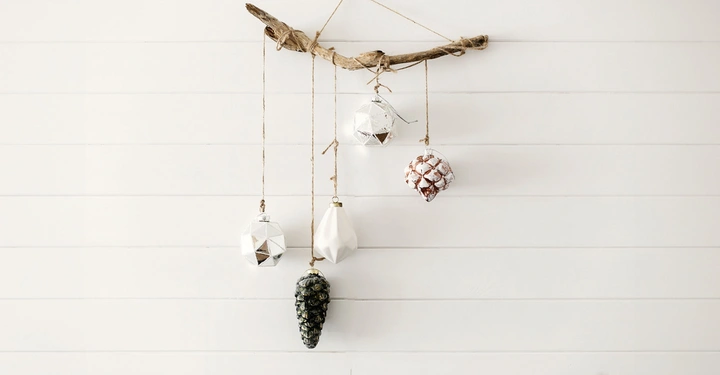 Astzweig hängt waagerecht an einer Schnur von der Wand mehrere dekorative Weihnachtsornamente baumeln daran in einem Raum mit weißen Holzwänden