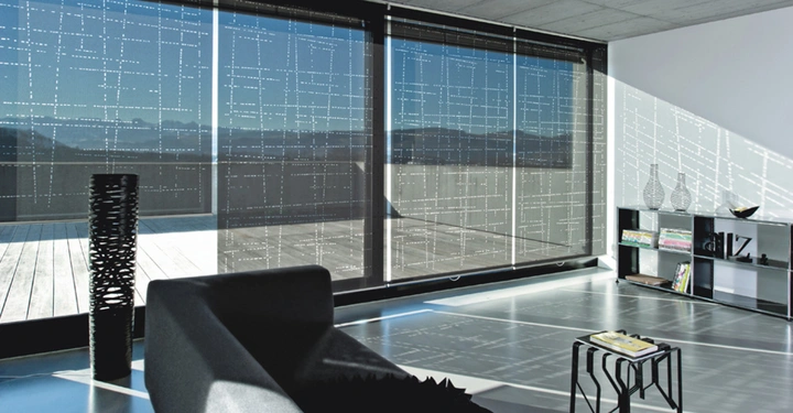 Große Fenster mit perforierten Jalousien lassen Licht in ein modernes Wohnzimmer mit grauem Sofa, kleinem Tisch und Regal vor einem Balkon herein.