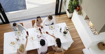 Sechs Personen sitzen an einem Esstisch und stoßen mit Weingläsern an in einem modernen Raum mit Holzfußboden und Blick auf eine Terrasse mit Wasser.