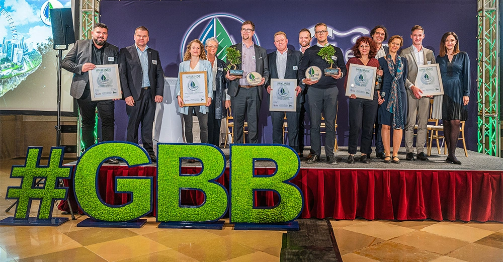 Eine Gruppe von Menschen hält Urkunden und Pflanzen steht auf einer Bühne in einem Konferenzraum Die Szenerie umfasst große leuchtende grüne Buchstaben #GBB. Text gefunden im Bild: - URKUNDE - KLIMAFIT - SEI DABEI! - LÖSEN WIR GEMEINSAM DAS KLIMAPROBLEM - ÖKOLAROTTENBACH