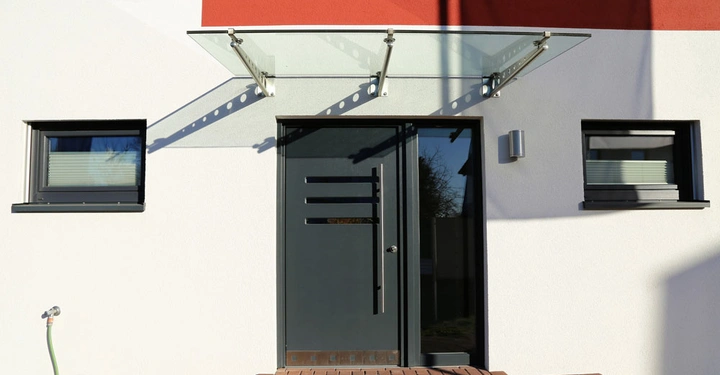 Eine moderne Haustür mit Glasvordach und zwei Fenstern befindet sich in einer weiß gestrichenen Hausfassade. Links ist ein angeschlossener Gartenschlauch sichtbar.
