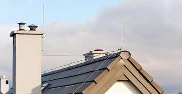 Ein Dach einer modernen Gebäudes mit einer Blitzschutzanlage bestehend aus Metallstäben und -drähten im Freien unter einem bewölkten Himmel.