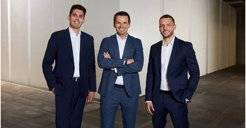 Drei Männer in dunkelblauen Businessanzügen ohne Krawatte stehen freundlich lächelnd in Firmenhalle.
