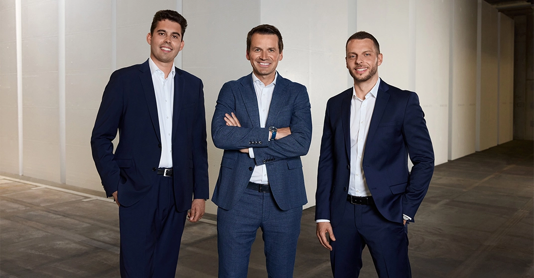 Drei Männer in dunkelblauen Businessanzügen ohne Krawatte stehen freundlich lächelnd in Firmenhalle.