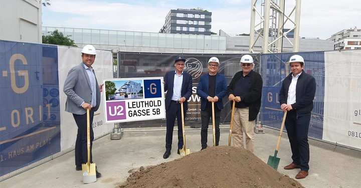 Fünf Männer tragen Helme und halten Schaufeln in den Händen Baustelle umgebend. Ein Mann hält ein Schild mit der Aufschrift U2 Leuthold Gasse 5B und einem Bild eines Gebäudes.