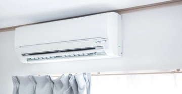 Klimaanlage hängt an einer weißen Wand bläst Luft in einen Raum darunter graue Vorhänge und ein Fenster mit Licht durchfluteten Vorhängen