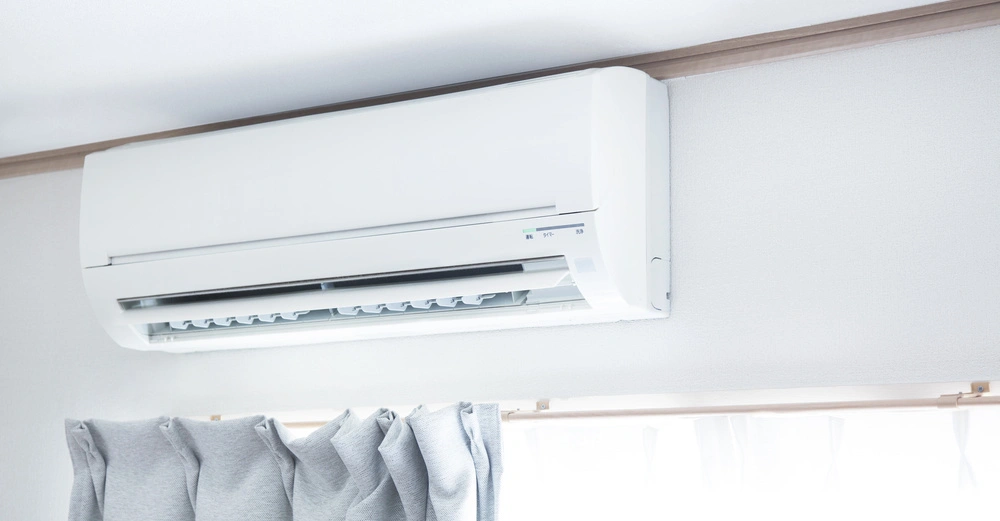 Klimaanlage hängt an einer weißen Wand bläst Luft in einen Raum darunter graue Vorhänge und ein Fenster mit Licht durchfluteten Vorhängen