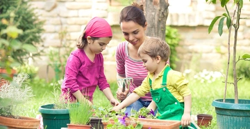 Zwei Kinder und eine Frau pflanzen zusammen Blumen in Töpfen in einem grünen Garten mit Steinmauer im Hintergrund.