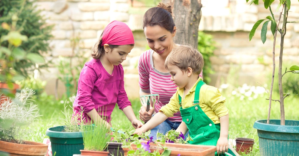 Zwei Kinder und eine Frau pflanzen zusammen Blumen in Töpfen in einem grünen Garten mit Steinmauer im Hintergrund.