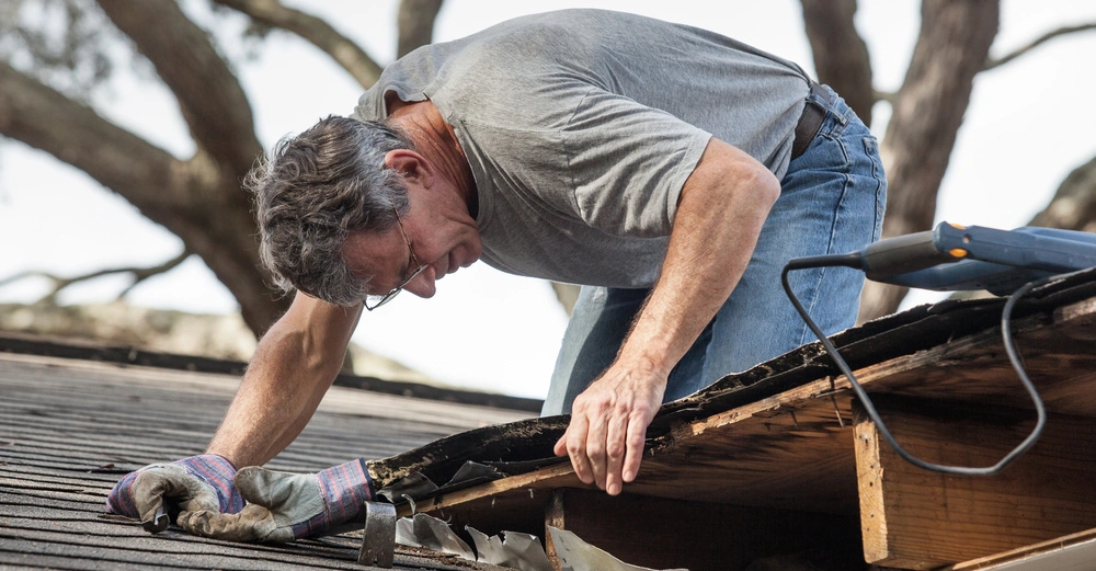 Mann in grauem T-Shirt und Jeans arbeitet auf einem Hausdach mit Handschuhen und einem Elektrowerkzeug, um Dachbeschädigungen zu reparieren Bäume im Hintergrund