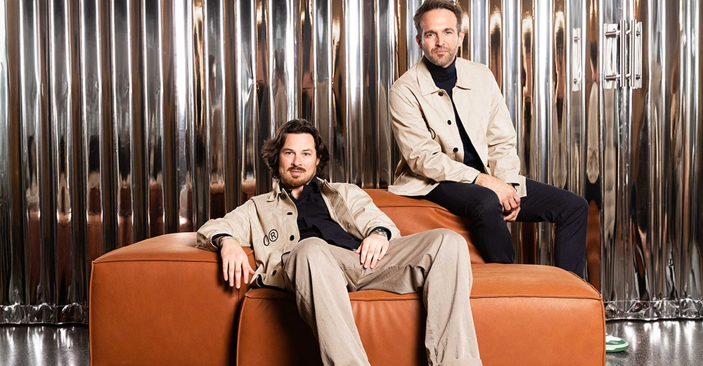 2 Männer mittleren Alters in beigen Anzügen bzw Sakkos gekleidet Einer sitzt in einem großen orangen Ledersessel, der andere rechts auf der Lehne. Im Hintergrund Wandbelag aus Metallstäben.