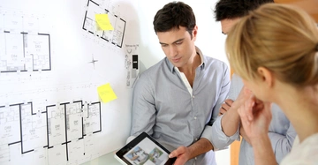 Drei Personen betrachten einen Tablet-Computer neben einem großen Architekturplan an der Wand eine Person zeigt auf das Tablet zwei gelbe Zettel stehen am Plan. Texte auf den Zetteln: Zettel 1 BEA + F05 Zettel 2 C+F + P.M.