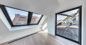 Dachgeschosszimmer mit Holzfußboden und großen Fenstern mit Blick auf Ziegeldächer und Terrasse Reihenhäuser und Himmel im Hintergrund.