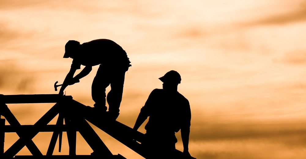 Zwei Personen arbeiten auf einem Dachrahmen beim Sonnenuntergang. Eine Person benutzt einen Hammer, während die andere daneben steht. Der Himmel im Hintergrund ist orangefarben.