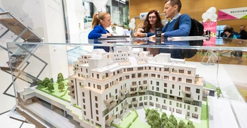 Modell eines modernen Mehrfamilienhauses in einer Glasvitrine Personen im Gespräch daneben heller Messekontext.