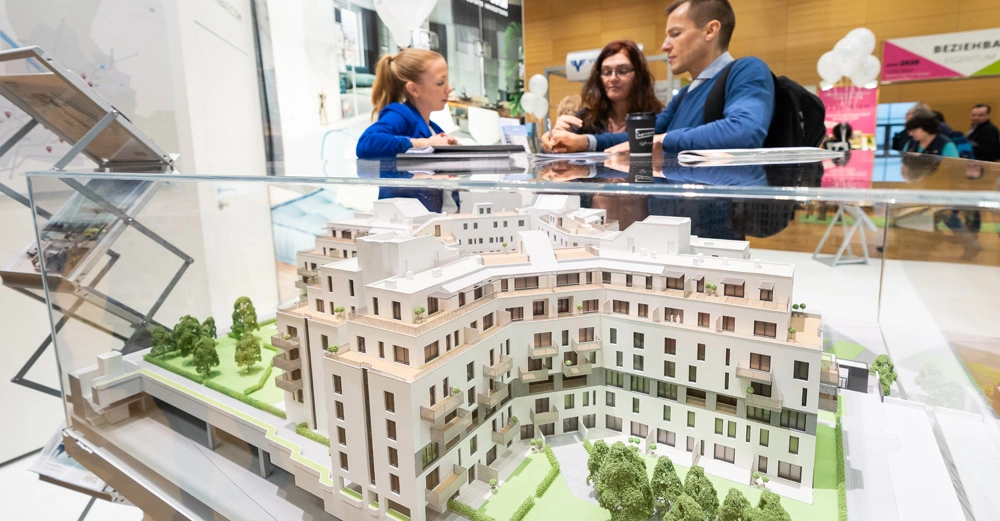 Modell eines modernen Mehrfamilienhauses in einer Glasvitrine Personen im Gespräch daneben heller Messekontext.