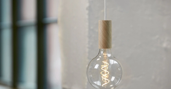 Galerie-Bild 6: Glühbirne hängt leuchtend an einer Schnur an Holzhalterung vor einer groben grauen Wand im Vordergrund mit einem unscharfen Fenster im Hintergrund