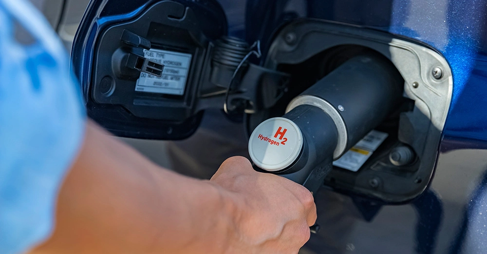 Ein Arm hält eine Wasserstoffpistole und tankt ein blaues Auto in einer Tankstelle. Text auf der Tankklappe Fuel Type Hydrogen.
